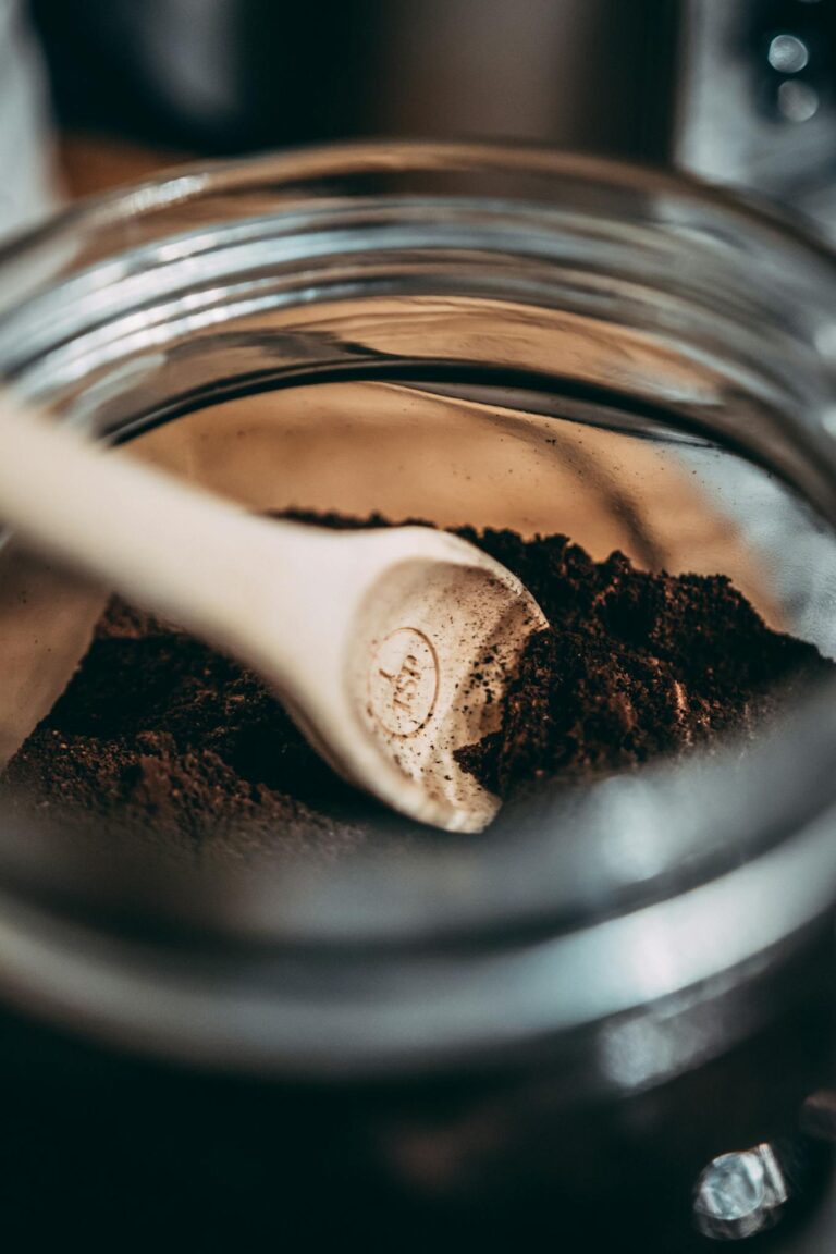 Wooden spoon resting in dark coffee grounds inside a glass jar.